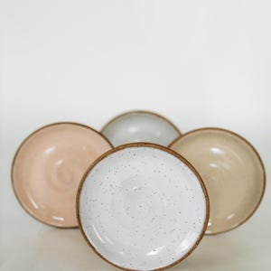 May include: A collection of ceramic plates in various colors, including white with speckled detailing, pale pink, beige, and gray. Each plate has a brown rim. The plates are arranged on a white background.