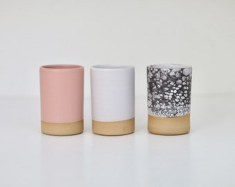 May include: Three ceramic cups with a speckled glaze. The cups are in shades of pink, white, and gray. The cups have a brown rim.