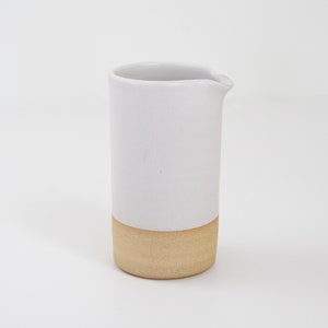 May include: A white ceramic pitcher with a beige bottom. The pitcher has a rounded lip and a smooth, matte finish.