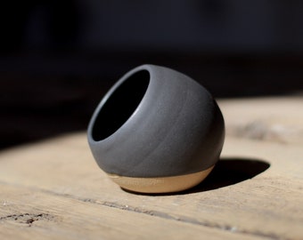 May include: A small, black ceramic bowl with a rounded shape and a slightly uneven surface. The bowl is tilted to the side, revealing a light brown interior.