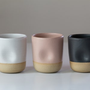 May include: Three ceramic mugs with a textured, uneven surface. The mugs are in white, light pink, and black with a light brown rim.