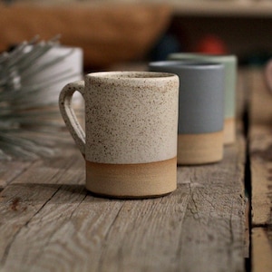 May include: A collection of ceramic mugs in various colors, including speckled beige, light blue, and pink, with a tan base. The mugs are arranged on a rustic wooden surface, showcasing their simple, modern design. Ideal for coffee or tea.