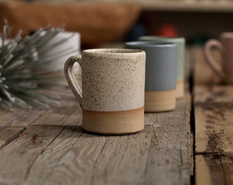 May include: Several ceramic mugs in a row, each with a different colour, including speckled beige, light blue, and pink, with a tan base. The mugs are on a rustic wooden surface, highlighting their simple, modern design. Suitable for coffee or tea.