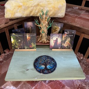 May include: A green wooden table with a tree of life design on a round plate. There are seven cards on the table with images of nature and the words "Dragon", "Whispering Pelican", "Wisdom of the Grove", "Wild Spirit", "Raven Spirit", "Rainbow Waterfall", and "Inner Spirit".