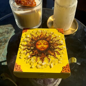 May include: A yellow wooden box with a sun design, surrounded by crystals, sits on a reflective surface. A candle with a large crystal and a selenite tower are also present. The box has ornate corner designs.