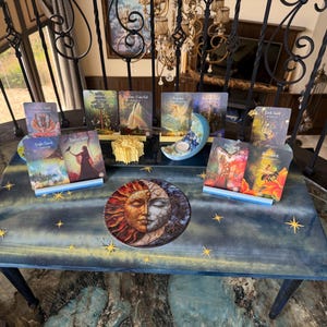 May include: A collection of oracle cards displayed on a blue table with a celestial design and gold star accents. The cards feature various illustrations and titles, including "Eagle Spirit" and "Earth Spirit." A decorative sun and moon plate is also present.