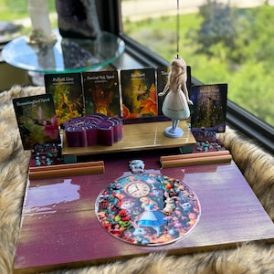 May include: A decorative display featuring an Alice in Wonderland theme. It includes a figurine of Alice, tarot cards, and a circular image with Alice and the White Rabbit. The display has a purple and gold color scheme.