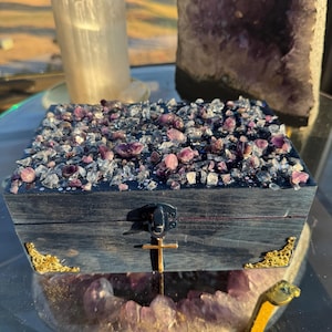 May include: A dark wooden box with a lid covered in small, sparkling crystals and gemstones. The box has a black clasp and gold-colored corner accents. The box is sitting on a glass surface with a large amethyst geode below.
