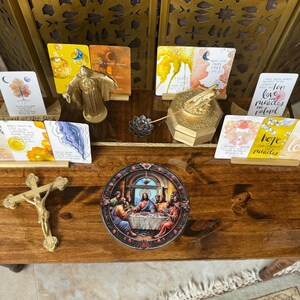 Puede incluir: Una mesa de madera con un crucifijo dorado, una pintura redonda de la Última Cena y varias tarjetas con mensajes inspiradores. Las tarjetas tienen fondos de acuarela y detalles dorados. La mesa tiene un acabado de madera marrón oscuro.