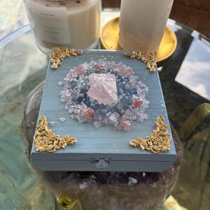 May include: A light blue wooden box with gold filigree corners, adorned with a central arrangement of crystals and a large pink quartz. A candle with a crystal top and a white crystal are also visible.