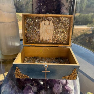 May include: Open wooden box with a blue exterior and gold accents. The interior is lined with glitter and a carved angel figure. The box is resting on a large amethyst geode, with a selenite tower in the background.
