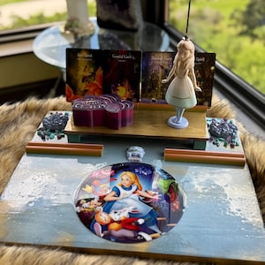 Puede incluir: Una exhibición decorativa con el tema de Alicia en el país de las maravillas. Incluye una imagen de Alicia recubierta de resina, una figura y tarjetas con texto. La exhibición tiene una base de madera con elementos decorativos y una caja morada.