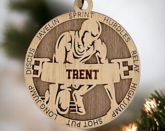 May include: Wooden ornament featuring a silhouette of a runner in a starting position. The ornament is decorated with the words "Sprint", "Hurdles", "Relay", "High Jump", "Long Jump", "Shot Put", "Javelin", "Discus" around the edge. The name "Trent" is inscribed on the ornament.