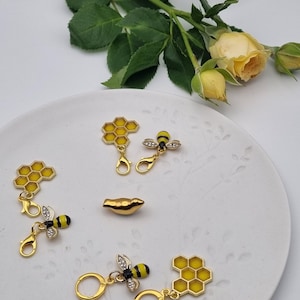 Könnte beinhalten: Eine Sammlung goldfarbener Bienen- und Wabenanhänger mit gelben und schwarzen Emaille-Details. Die Anhänger werden auf einem weißen Teller mit floralem Design zusammen mit einer goldfarbenen Perle und gelben Rosen präsentiert.