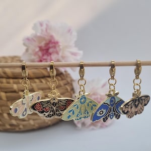 Könnte beinhalten: Fünf goldfarbene Mottenanhänger mit aufwendigen Emaille-Designs in verschiedenen Farben, darunter Rosa, Schwarz, Blau und Beige. Jeder Anhänger ist an einem goldenen Verschluss befestigt. Die Anhänger werden vor einem verschwommenen Hintergrund mit Blumen und einem geflochtenen Korb präsentiert.