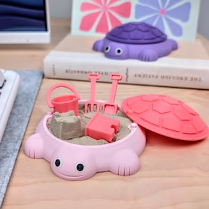 以下が含まれることがあります： 砂が入ったライトピンクの亀の形をした砂場容器。小さなピンクのバケツ、ピンクのシャベル、ピンクの熊手、ピンクの四角い型が含まれています。お揃いのピンクの亀の甲羅の蓋が横に置かれ、本の上には紫色の亀のおもちゃが乗っています。