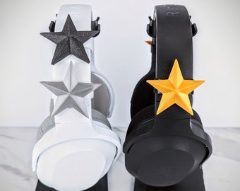 Può includere: Due supporti per cuffie, uno nero e uno bianco, con decorazioni a forma di stella. Il supporto nero ha una stella dorata, e il supporto bianco ha una stella argentata e una stella nera.