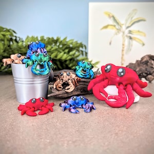 May include: A collection of seven small, colorful, hand-painted crab figurines. The crabs are made of a smooth, glossy material and have realistic details, including eyes, claws, and legs. The crabs are arranged on a wooden surface and a metal bucket.