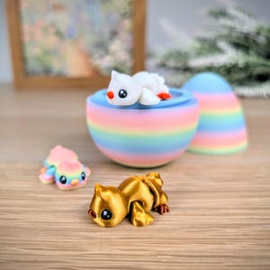 May include: A collection of colorful, whimsical figurines. A gold-colored cat figurine is in the foreground. A rainbow-striped egg-shaped container holds a white seal figurine. A smaller, pastel-colored seal figurine is also present.