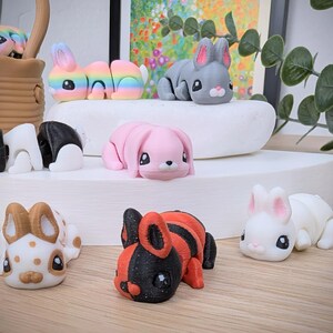 May include: A selection of 3D-printed rabbit figurines in a variety of colours. The rabbits are in shades of rainbow, pink, grey, white, and black with orange stripes. Each rabbit has large eyes and a rounded shape.
