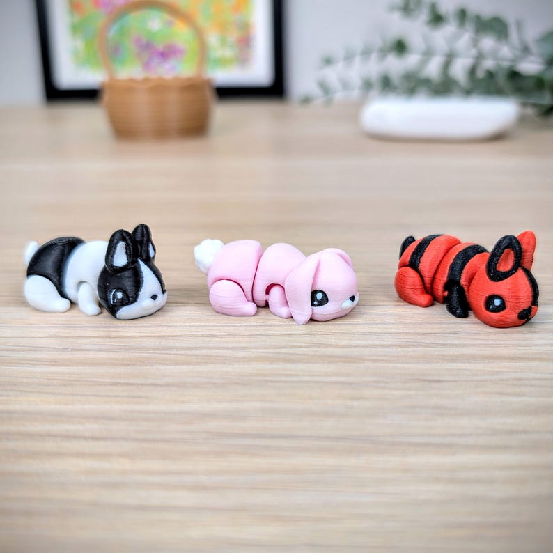 May include: Three small, decorative bunny figurines. One is black and white, one is pink, and the third is red and black. The bunnies are lying down on a light brown wooden surface. The background includes a framed picture and a small basket.