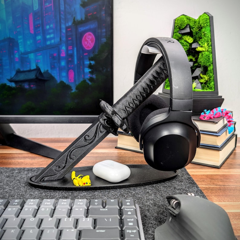 May include: A black headphone stand shaped like a katana sword, holding a pair of black headphones. The stand has a circular base with a small yellow figurine and a white case. A computer monitor and keyboard are in the background.