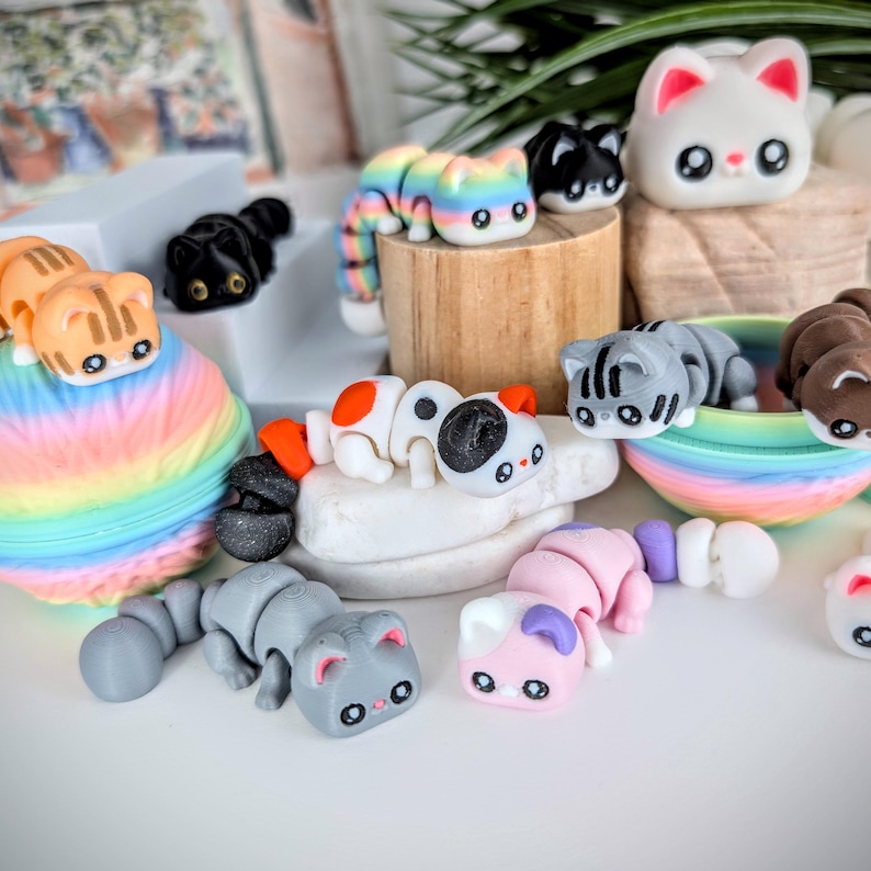 May include: A collection of colorful, miniature cat figurines. The cats are in various colors and patterns, including rainbow, black, gray, and white. Some are lying down, while others are standing. The figurines are displayed on a white surface.