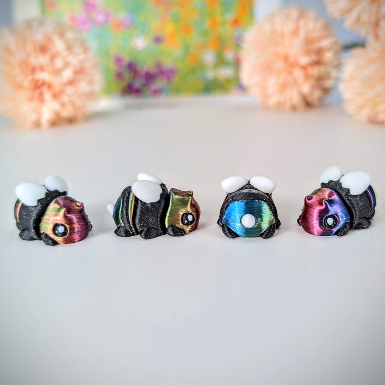 May include: Four small, iridescent, 3D printed bumblebees in black, blue, green, and red. The bumblebees are facing forward with their wings spread out.