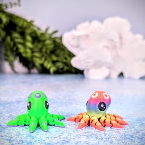 May include: Two small octopus figurines, one green with purple accents and the other with rainbow colors. The octopus figurines are on a light blue surface.