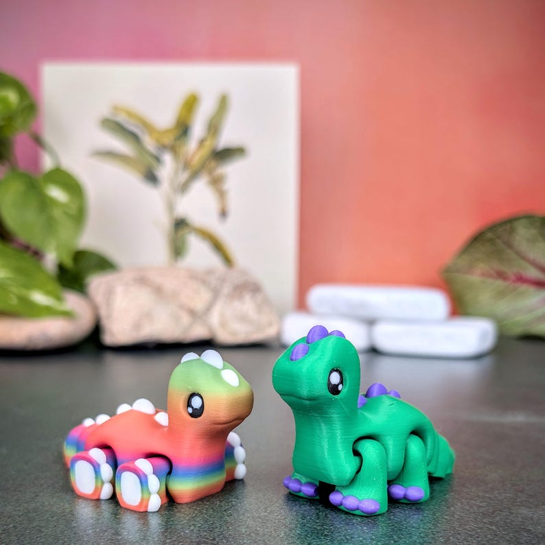 Op de afbeelding: Twee kleine, kleurrijke dinosaurusbeeldjes. E&eacute;n is rood, oranje, geel en groen met regenboogstrepen. De andere is groen met paarse accenten. Beide hebben witte details en grote ogen.
