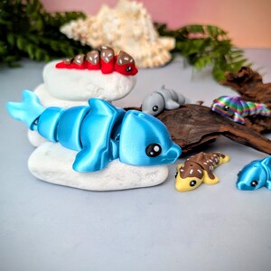 May include: A collection of colorful, articulated dolphin figurines. The main dolphin is bright blue, resting on a white stone. Other dolphins are gray, yellow, brown, and rainbow-colored. The background includes driftwood and greenery.
