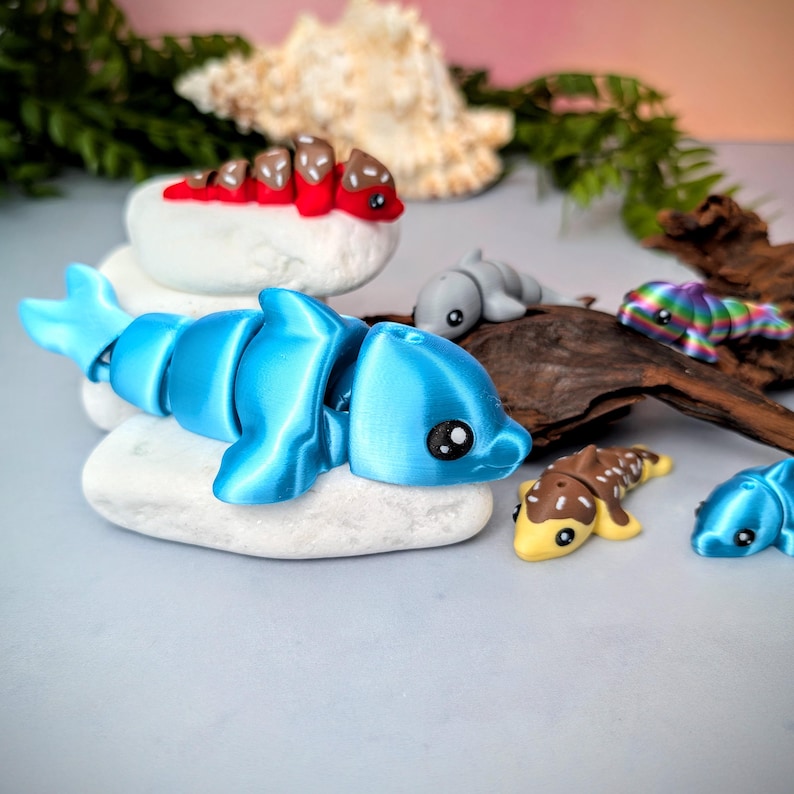 May include: A collection of colorful, articulated dolphin figurines. The main dolphin is bright blue, resting on a white stone. Other dolphins are gray, yellow, brown, and rainbow-colored. The background includes driftwood and greenery.