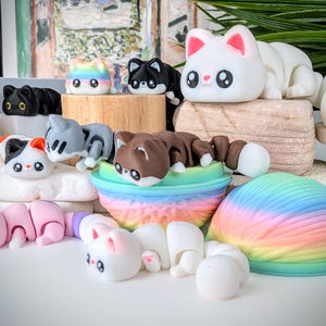 May include: A collection of colorful, articulated cat figurines. The cats are in various colors, including black, white, gray, and brown, with rainbow and pastel accents. Each cat has large, expressive eyes and is posed in a playful manner. The figurines are displayed on a white surface.