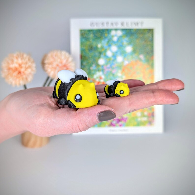 May include: Two yellow and black bee figurines, one larger than the other, are held in a hand. The larger bee has a white face with black eyes.