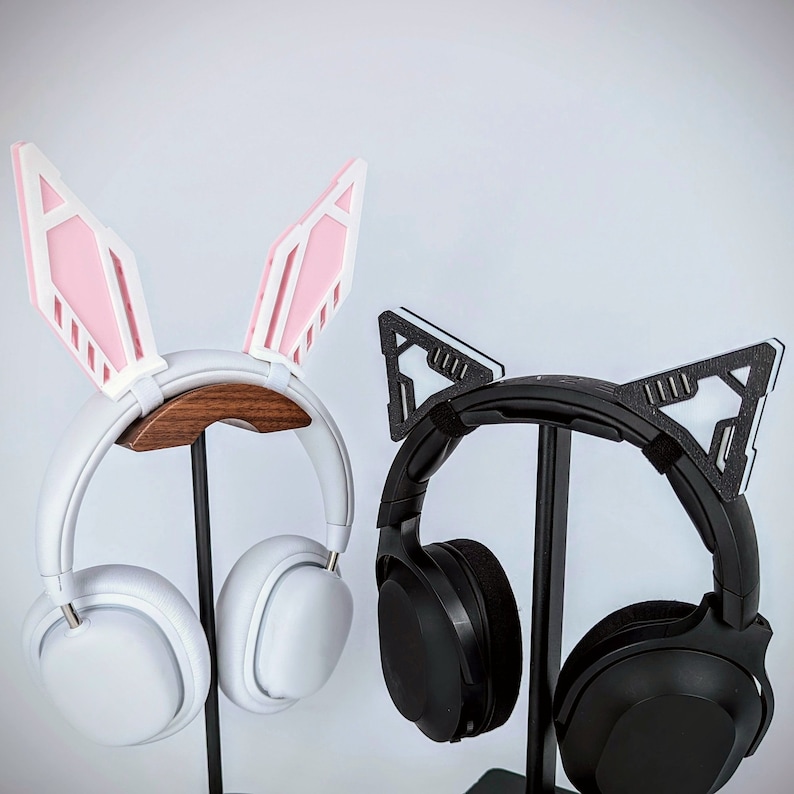 Pode incluir: Dois conjuntos de fones de ouvido com acess&oacute;rios decorativos para as orelhas. Um conjunto &eacute; branco com orelhas de coelho rosa e branco, o outro &eacute; preto com orelhas de gato pretas e brancas. Ambos os conjuntos est&atilde;o em suportes pretos sobre um fundo branco.