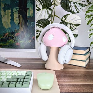 May include: White headphones with a pink mushroom-shaped stand. The stand has a white top with two small circles and a white base. The headphones are on a wooden desk with a green keyboard and a green mouse.