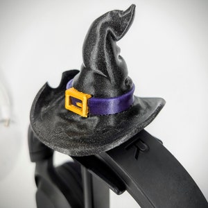 May include: A black witch hat with a purple and gold buckle sits on top of a black headphone headband. The headband has the letter "R" printed on it.