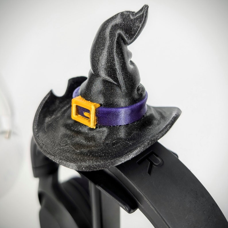 May include: A black witch hat with a purple and gold buckle sits on top of a black headphone headband. The headband has the letter "R" printed on it.