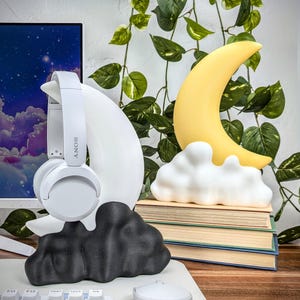 Pode incluir: Auscultadores brancos com a palavra "SONY" impressa no lado, apoiados num suporte branco em forma de lua crescente. O suporte est&aacute; sobre uma base preta em forma de nuvem. Uma luz noturna amarela em forma de lua crescente est&aacute; sobre uma pilha de livros.
