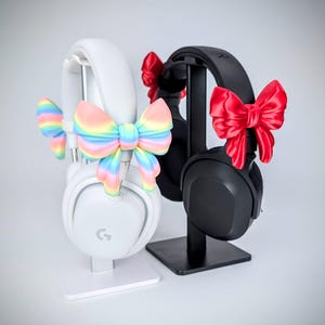 May include: Two sets of headphones, one white and one black, displayed on stands. The white headphones are adorned with a pastel rainbow bow, while the black headphones feature a red bow. The headphones are a modern design.