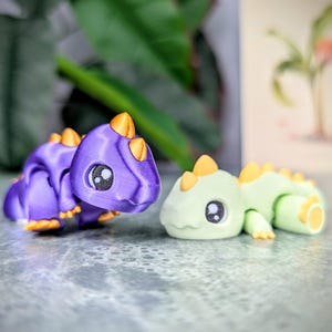 May include: Two 3D-printed toy dinosaurs. One is purple with orange accents, the other is light green with yellow accents. Both have large, round eyes and are posed on a textured surface. The toys are likely designed for children.