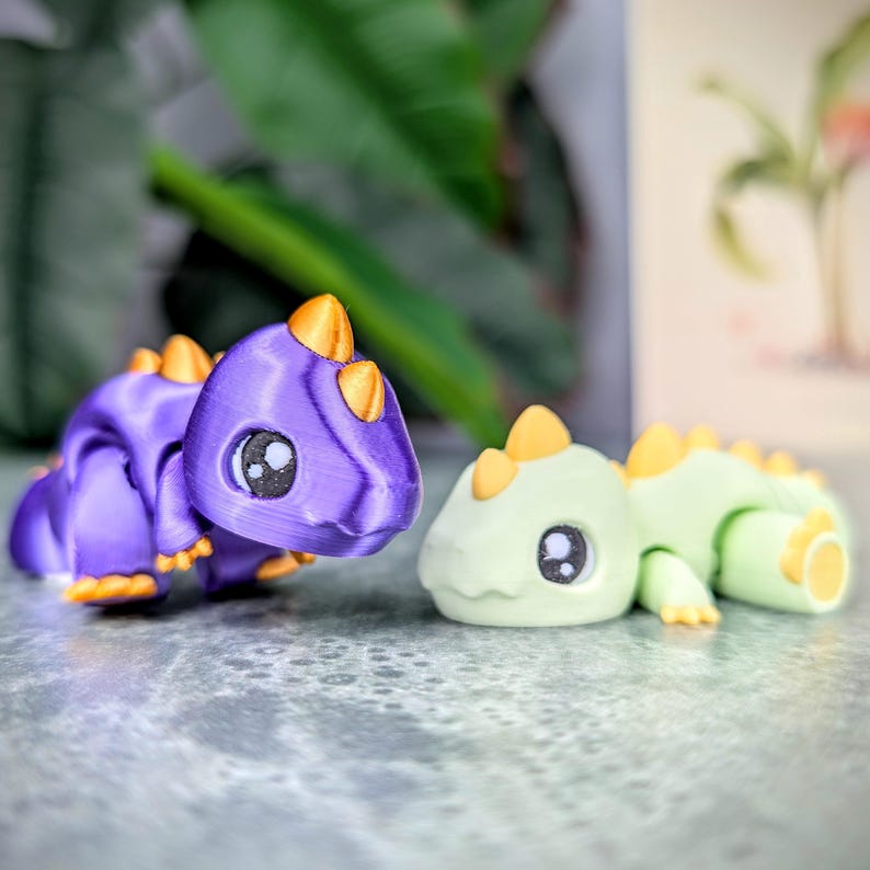 May include: Two 3D-printed toy dinosaurs. One is purple with orange accents, the other is light green with yellow accents. Both have large, round eyes and are posed on a textured surface. The toys are likely designed for children.