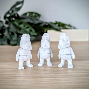 May include: Three white figurines with a speckled texture, standing in a row on a wooden surface.