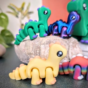Op de afbeelding: Een verzameling kleurrijke, beweegbare dinosaurusfiguren. De dinosaurussen zijn in tinten geel, groen, blauw en rood, met contrasterende accenten. De figuren zijn geplaatst op een stenen oppervlak, waarbij hun ontwerp en details worden getoond.