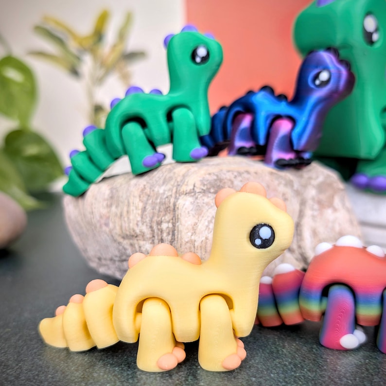 Op de afbeelding: Een verzameling kleurrijke, beweegbare dinosaurusfiguren. De dinosaurussen zijn in tinten geel, groen, blauw en rood, met contrasterende accenten. De figuren zijn geplaatst op een stenen oppervlak, waarbij hun ontwerp en details worden getoond.