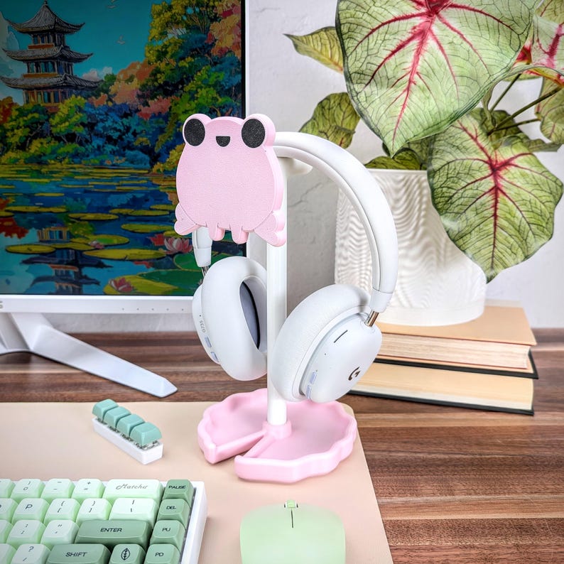 Pu&ograve; includere: Supporto per cuffie a forma di rana rosa con cuffie bianche. Il supporto ha una base rosa e un palo bianco. Le cuffie sono appoggiate sul supporto.