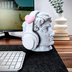 May include: White headphones with a pink heart on the headband, resting on a white bust sculpture of a man's head. The bust is on a black surface with a white keyboard in the foreground.