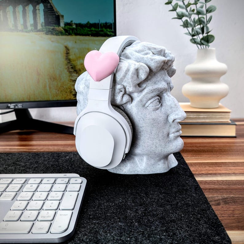May include: White headphones with a pink heart on the headband, resting on a white bust sculpture of a man's head. The bust is on a black surface with a white keyboard in the foreground.