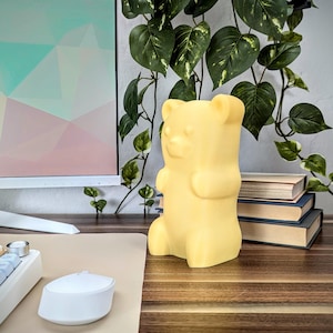May include: A yellow gummy bear-shaped night light on a wooden surface. The night light is in front of a computer monitor and a potted plant.