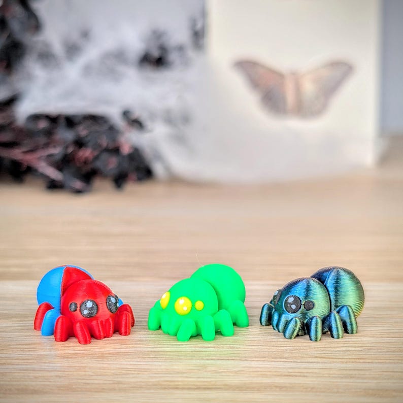 May include: Three colorful, stylized spider figurines. One is red and blue, another is green with yellow spots, and the third is blue and green. The spiders have multiple legs and large eyes, and are set against a blurred background.